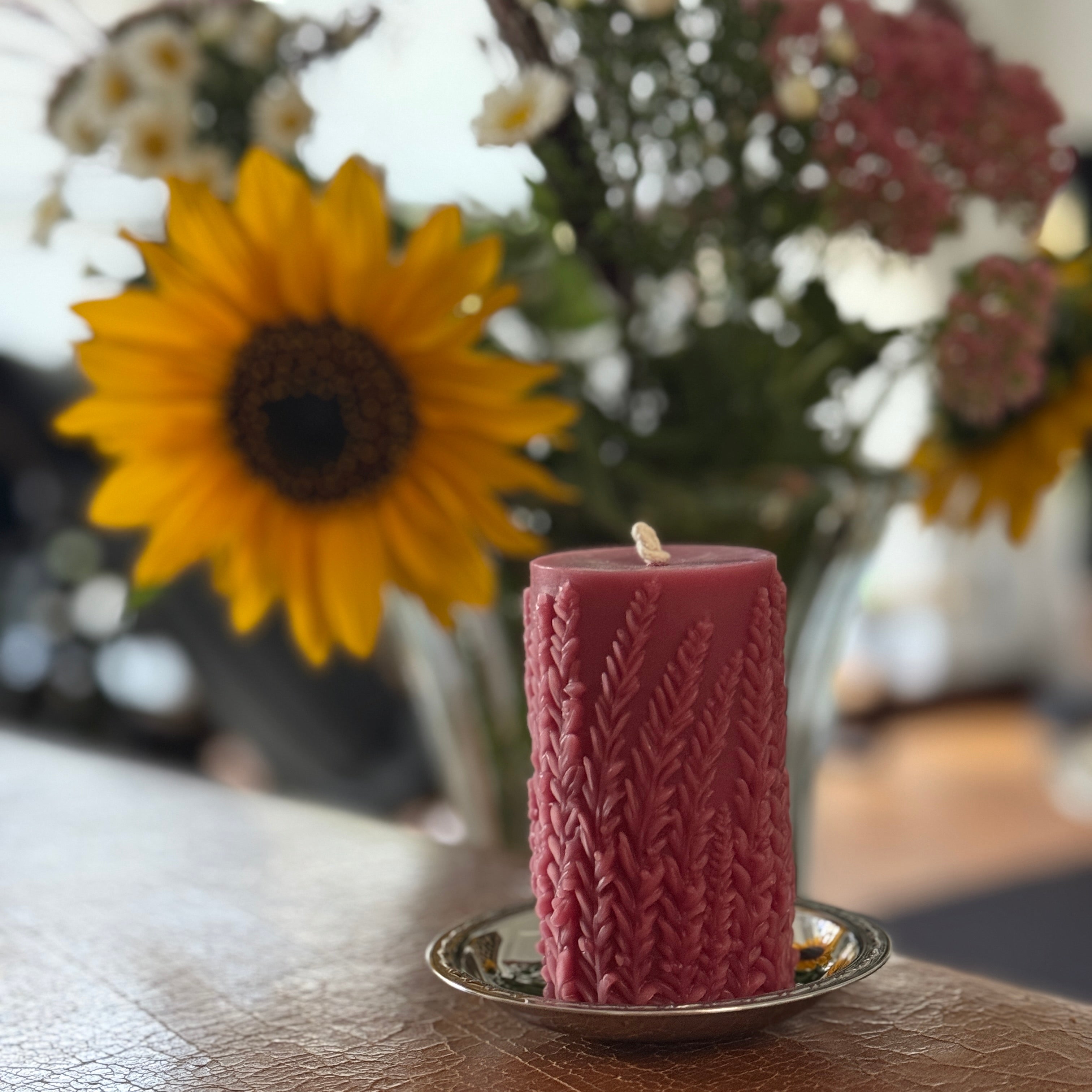 Eine Kerze mit Pflanzenrelief aus Bienenwachs in rot vor einem Blumenstrauch mit Sonnenblumen.