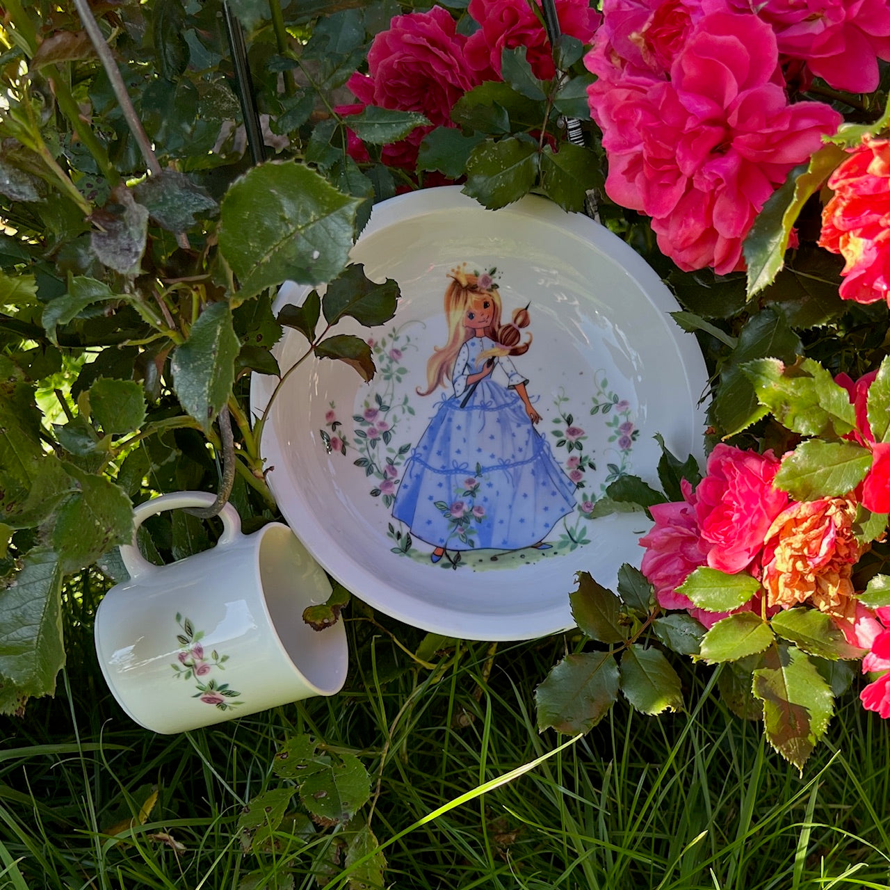 Ein Teller und eine Tasse mit Dornröschen darauf unter einem Rosenstrauch.
