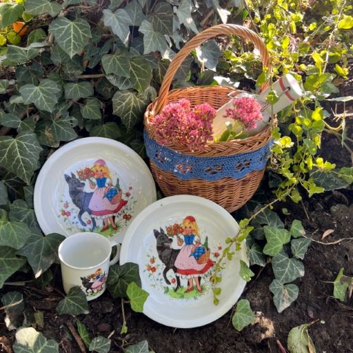 Zwei Teller und eine Tasse mit Rotkäppchen darauf neben einem Korb im Wald.