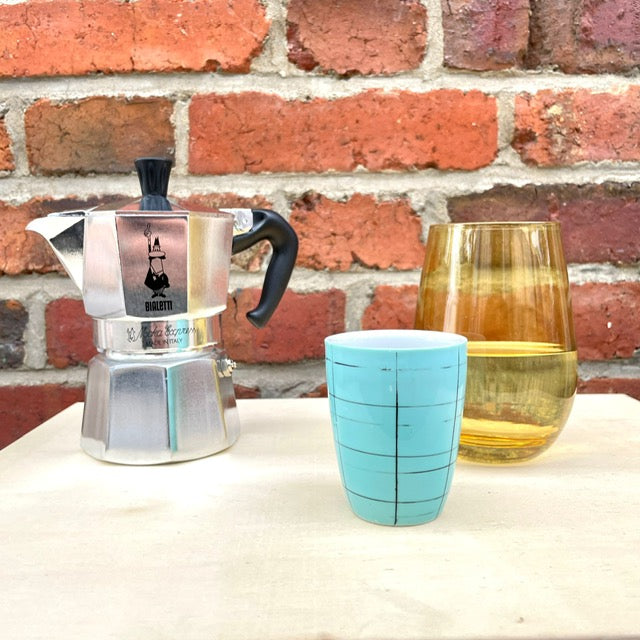 Bialetti in Alu mit einem Wasserglas und einer Espressotasse vor einer ziegelwand. 