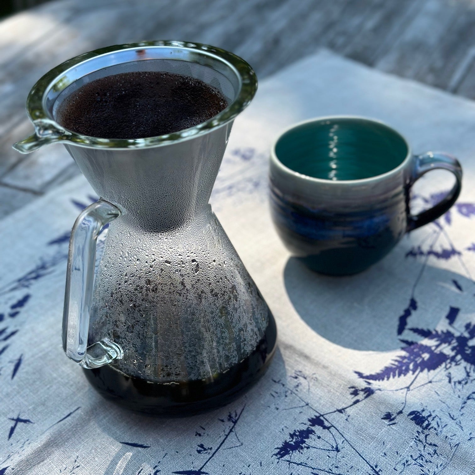 kaffeebereiter aus glas mit edelstahlfilter auf dem tisch mit blauer keramiktasse