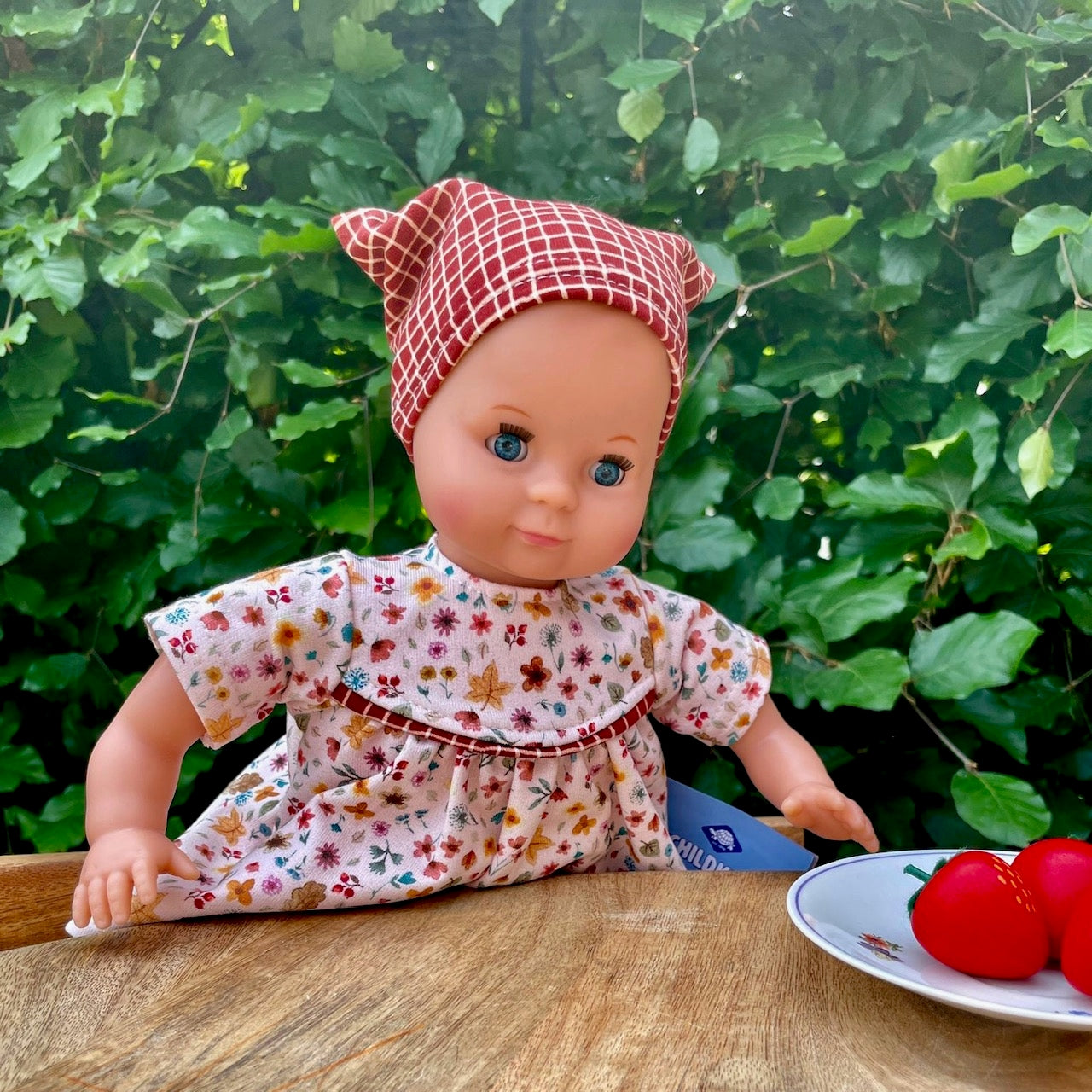 puppe am tisch sitzend mit rotem kopftuch und gebluemten kleid.
