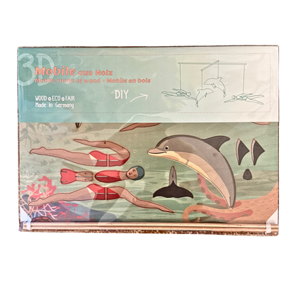 Ein Mobile Bastelset aus Holz mit zwei Schwimmerinnen und zwei Delfinen. Verpackung in Folie. 