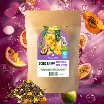 Papiertüte mit Eistee Mango. Im Hintergrund zerschnittene Mangos und davor die Tee-Kräuter.