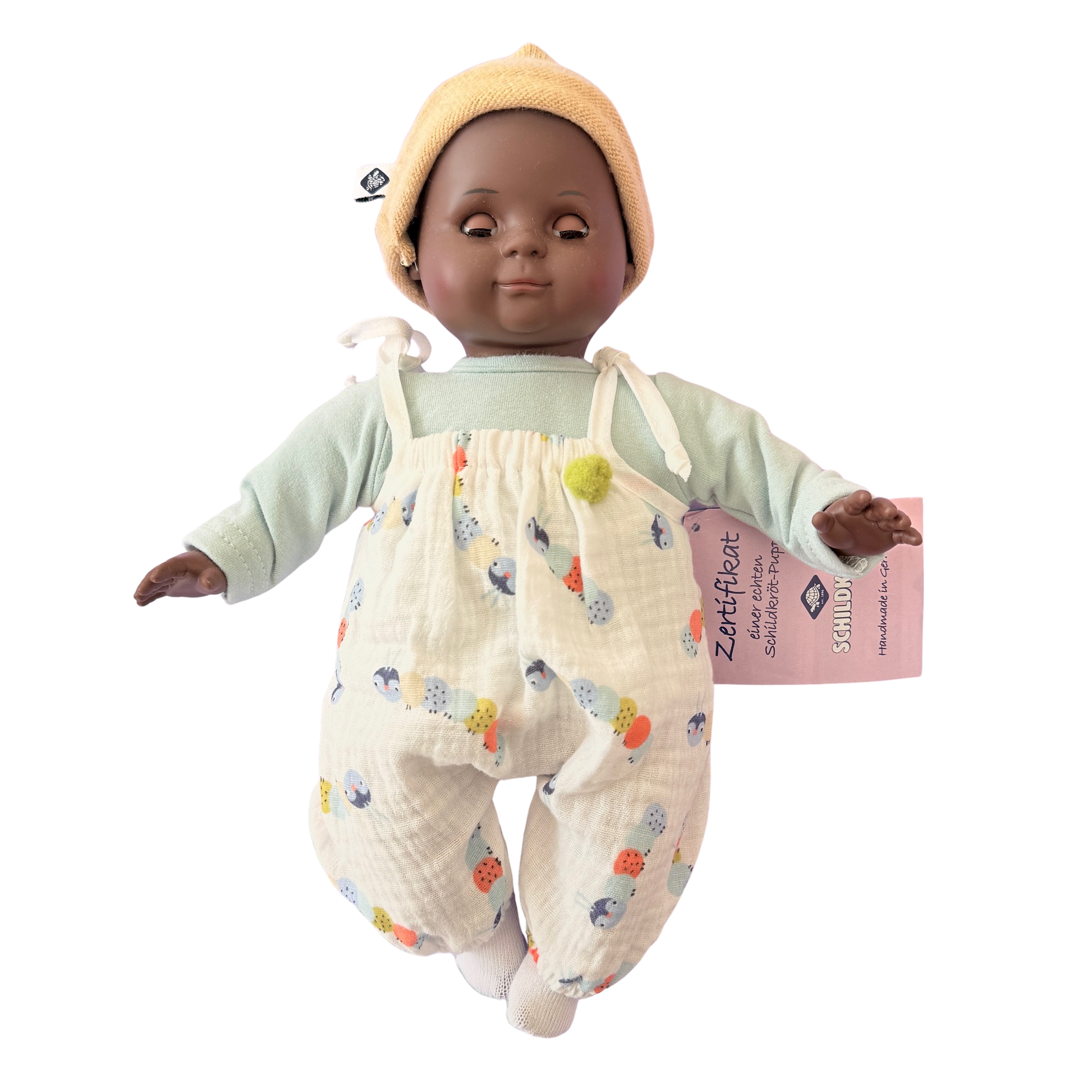 Kleine farbige Puppe mit Schlafaugen und Stoffkörper. Ein weißer Strampler und ein Mützchen. 