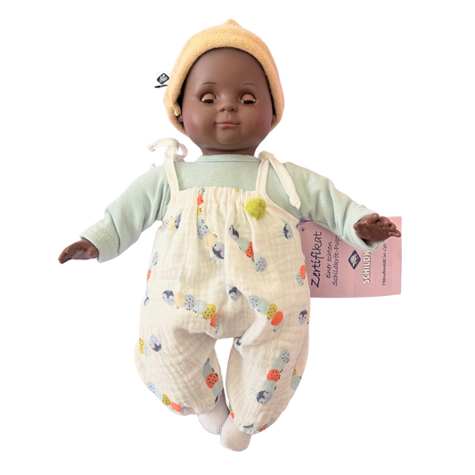 Kleine farbige Puppe mit Schlafaugen und Stoffkörper. Ein weißer Strampler und ein Mützchen. 