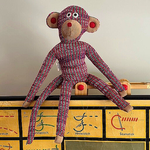 Kuscheltier Affe mit einem langen Schwanu und großen Ohren aus gestrickter rot-beiger Baumwolle sitzend auf einem Schulposter